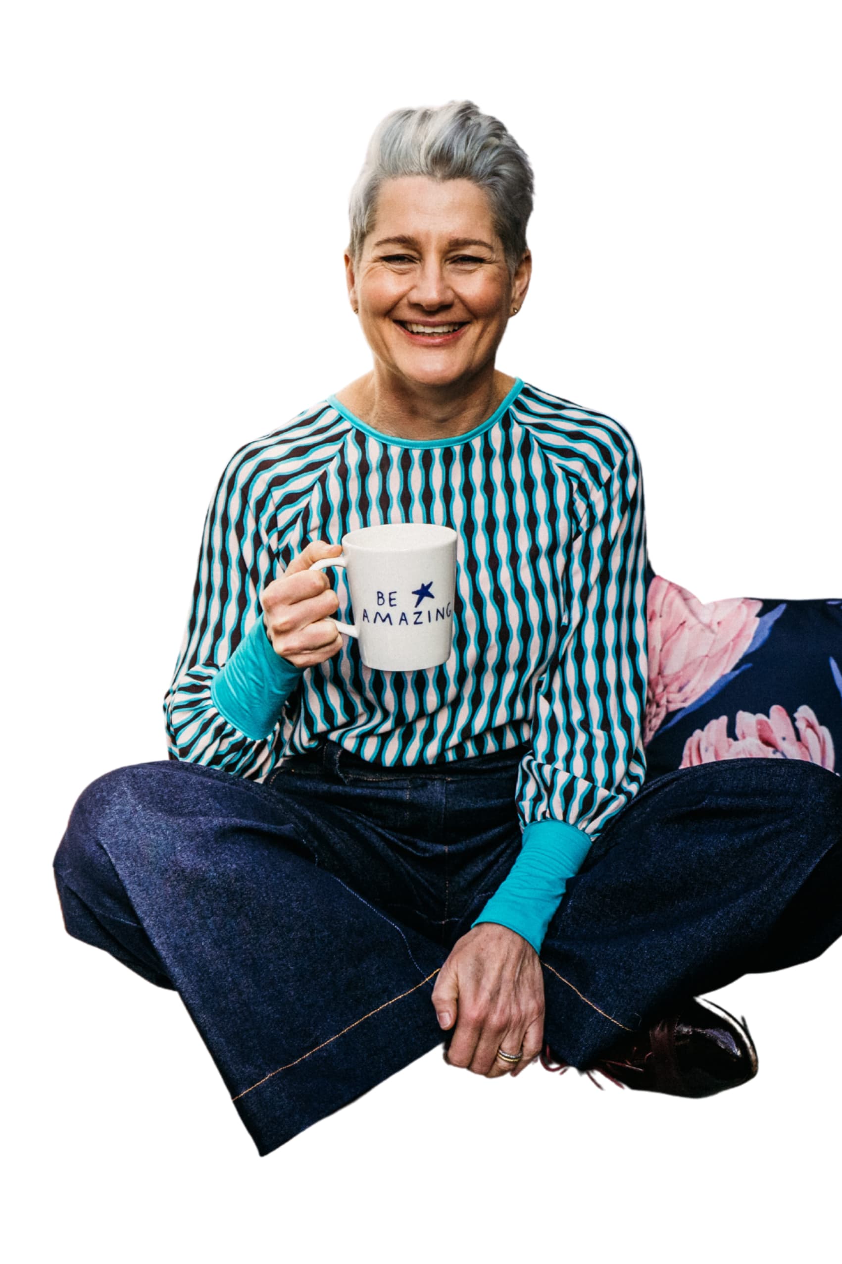 A lady with grey hair is sitting cross-legged and smiling at the camera. She is wearing a turquoise top and holding a mug that reads be amazing.